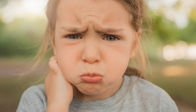 Close up portrait of a sad young child pouting with a grumpy facial expression and furrowed eyebrows while holding a hand to the cheek outdoors