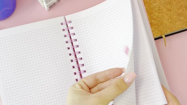 Young woman's manicured hands placing a pink bookmark on a blank page of a gridded notebook and then flipping through the pages, a concept of studying and organization