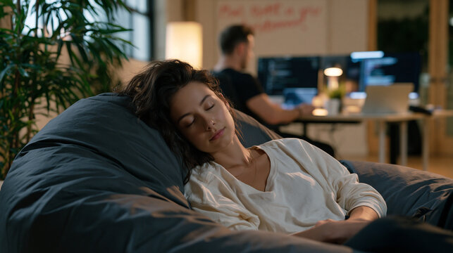 Young founder collapsed asleep on a beanbag in a tiny office while her business partner still codes at a standing desk behind her, a motivational whiteboard message half-erased, the imbalance of res