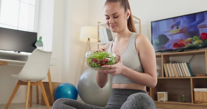 Woman athlete eating salad at home. Fit female enjoys fresh greens after workout in cozy room, focusing on healthy nutrition, mindfulness, wellness, and fitness. Pure healthy life concept here