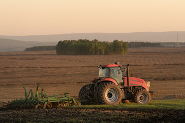 Fototapeta premium Red tractor on vast agricultural land among rolling hills and farmland at sunset