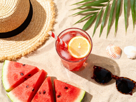 Refreshing beach picnic with watermelon slices, lemonade jar, sunglasses, straw hat, and seashells on white fabric