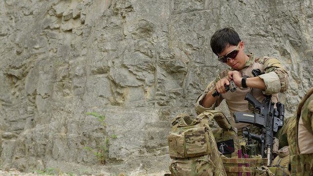 A focused military operator prepares his tactical gear, loading a magazine against a rugged cliff face to ensure operational readiness for a mission.