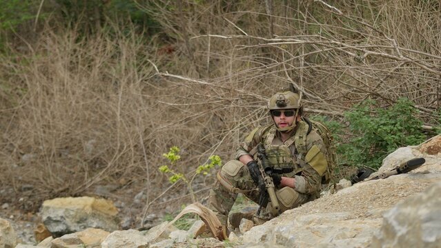 A special forces operator in full combat gear maintains a tactical overwatch position on a rugged, rocky hillside during a covert military operation.