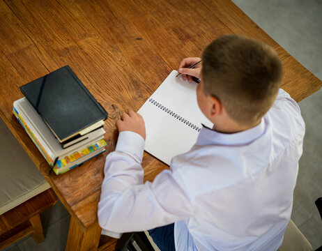 Top view of a schoolboy writing in a notebook beside a stack of books on a wooden table. Education, study planning, learning process and academic productivity concept.