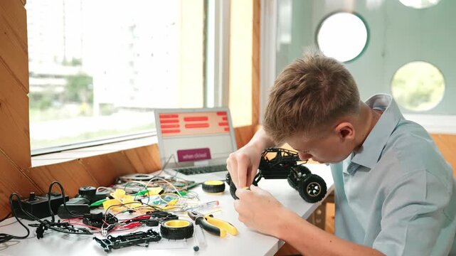Happy teenager fixing robotic model while using electronic equipment remove car wheel with wire and laptop scatter around. Smart boy study or learn about car robot construction at class. Edification