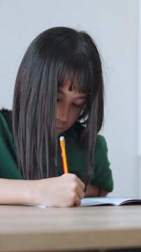 Vertical video showing close up selective focus of a focused asian kid girl writing in a notebook in daylight. Ideal for mobile social media, representing student doing homework and modern education.