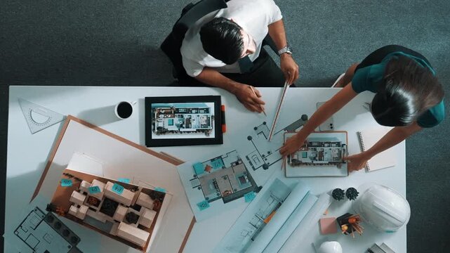 Top view of skilled businessman walking and give house design. Aerial view of architect engineer looking at blueprint and building structure at table with safety helmet and house model. Alimentation.
