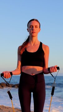 Attractive woman working out on the beach with resistance bands at sunset. Slow Motion. Vertical video.