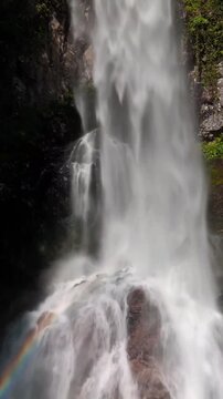 A stunning, powerful waterfall cascades down dark, mossy rock faces into a misty pool below, where a vibrant, beautiful rainbow arches through the rushing water spray.
 waterfall in the forest