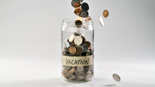 Close-up shot of coins falling into a coin jar labeled 'Vacation' in slow motion.
