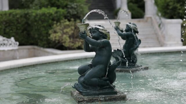 Ornamental cherub fountain statue surrounded by flowing water in an elegant garden setting. Classical sculpture and tranquil outdoor atmosphere.