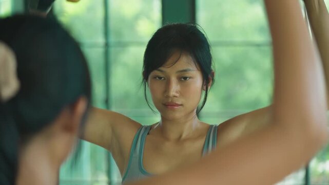 Asian woman in gray sportswear does a lateral raise with yellow dumbbells while facing a gym mirror with green trees outside.