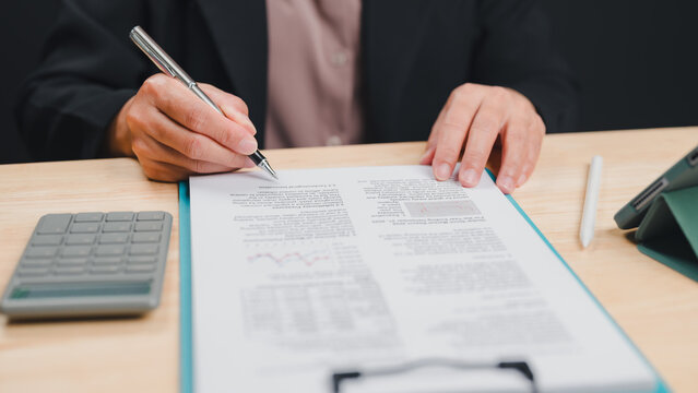 Professional business person wearing dark blazer sits wooden desk reviewing financial document clipboard holding silver pen near calculator tablet stylus workday office setting indoors this afternoon.