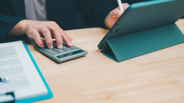 Professional accountant uses calculator alongside digital tablet including stylus pen analyzing financial document data atop wooden office desk daytime business hours performing tax preparation plus.
