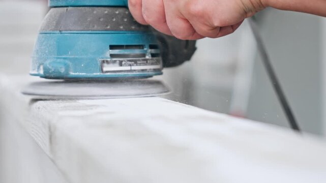 Close up of orbital sander head smoothing white painted wood plank, sanding dust flying in sunlight