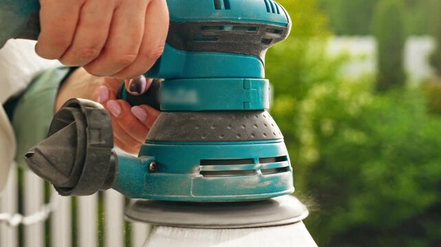 Woman with manicured hands using electric sander on white painted wooden railing in garden, close up