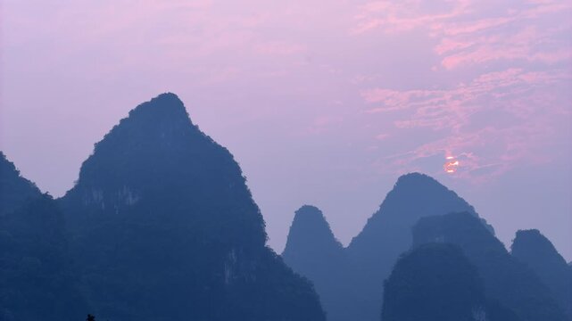 Timelapse of a beautiful pink and purple sunset, with the sun slowly setting behind the dramatic silhouettes of karst mountain peaks in Yangshuo, Guilin, China. Atmospheric evening twilight and golden