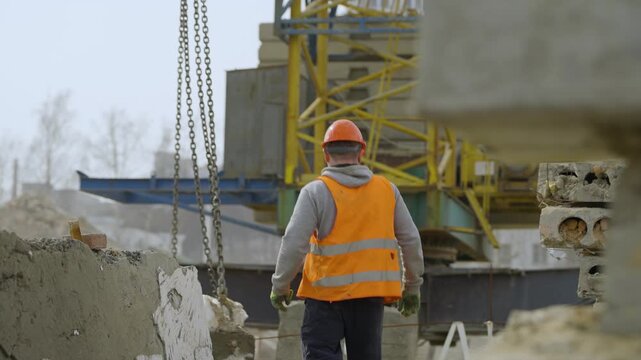 A construction worker walks on a construction site while lifting a beam with a crane