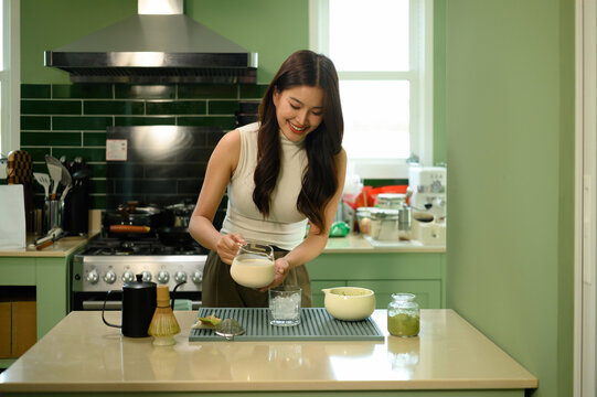 Asian Woman Making Matcha Latte at Home Kitchen Counter with Whisk and Matcha Powder Jar