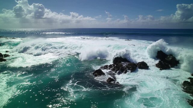 Drove view of turbulent ocean waves crashing against rocks with white foam and a blue sky