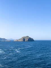 Fototapeta premium A scenic view of a rugged, rocky islet surrounded by deep blue sea water under a vast, clear blue sky with a faint moon visible during the day.