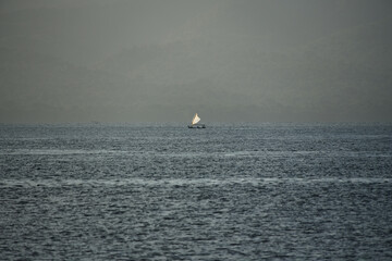 Minimalist long shot of a small traditional sailboat drifting on the vast calm ocean with a hazy, misty mountain horizon backdrop. © EkoPriyatno