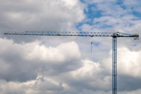 A tall blue tower crane with a long horizontal jib and yellow hook block photographed against a dramatic cloudy sky
