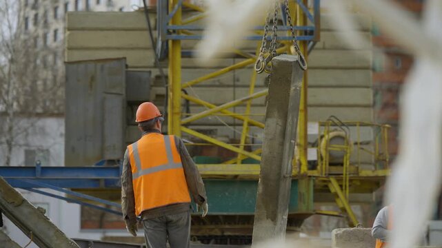 A slinger worker in an orange vest and helmet looks at the lifting of the beam by crane