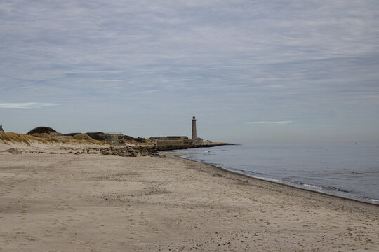 A lonely lighthouse stands at the edge of a sandy beach under a cloudy Nordic sky