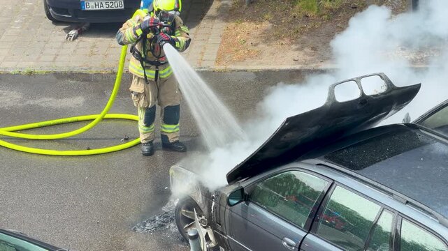 A firefighter blasts water into a smoking car engine compartment on a city road. Dense steam, soot and open panels show the fire being suppressed.