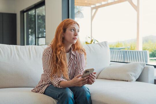 Redhead woman sits on a comfortable sofa holding her smartphone, looking thoughtfully into the distance, perhaps waiting or contemplating while relaxing at home