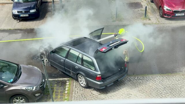 A firefighter sprays water across the street toward a smoke filled burned car. The open tailgate, hoses and urban parking area frame the emergency operation.