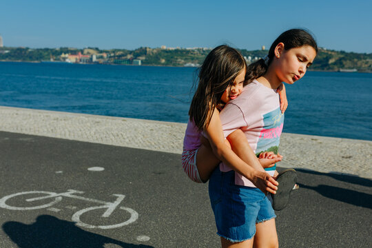 Older Sister Carrying Younger Sister Along a Waterfront Promenade