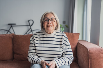 Obraz premium Senior woman with grey hair and glasses sits comfortably on a brown sofa at home, wearing a striped sweater and smiling warmly at the camera in a cozy living room portrait