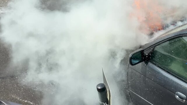 Heavy smoke and water spray surround the charred front wheel of a burned car. Soot, foam and wet asphalt reveal severe damage from the vehicle fire.