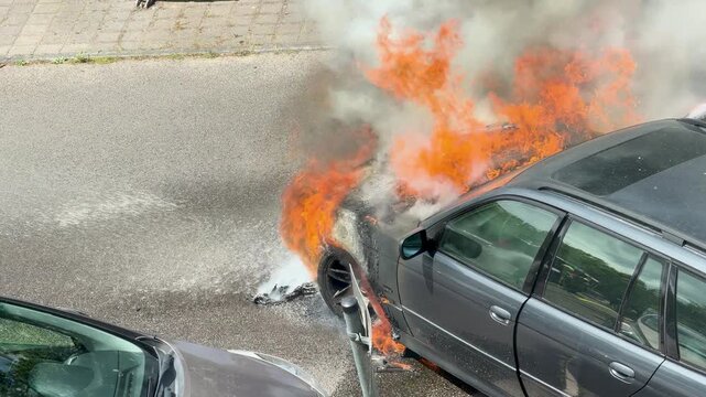 Large orange flames engulf the front of a parked station wagon before suppression. Black smoke, burning debris and heat create a dramatic urban car fire scene.