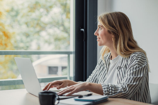 Young woman working from home on a laptop, pausing to smile and gaze out the window as she plans tasks, balances work and life, and finds calm inspiration in natural light