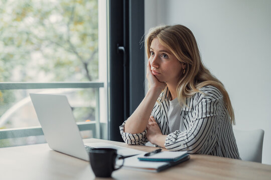 Young woman at a desk with laptop, staring out the window, showing boredom, fatigue and procrastination while working from her home office, unmotivated and pensive