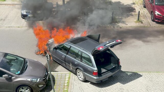 A station wagon burns on a city street with the rear hatch open. Black smoke, orange flames and parked cars create a dramatic emergency scene.