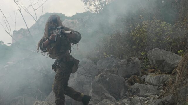 A special forces soldier in a ghillie suit advances through a dense smoke screen on a rocky mountain, conducting a covert tactical military operation.