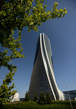 The striking, twisted glass facade of the Generali Tower (Torre Hadid) rises into a clear blue sky, framed by lush green ginkgo biloba tree leaves in the CityLife district.