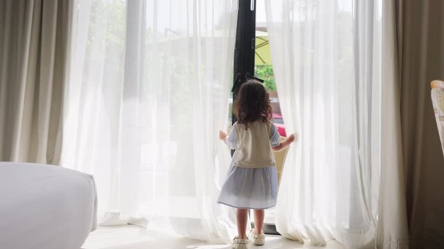 A curious young girl peeks through sheer white curtains by a window at home, looking at the camera with a gentle sense of discovery and childhood wonder.