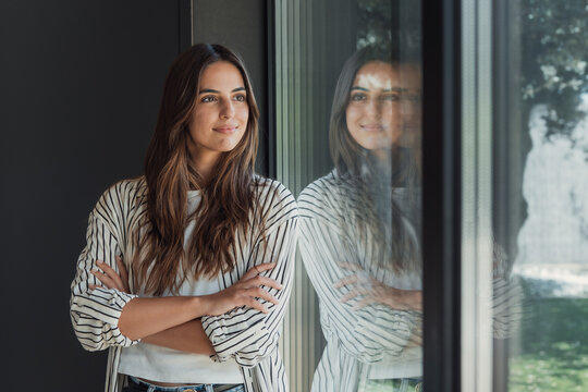 Woman standing with crossed arms, looking out a large window and reflecting on her thoughts, her reflection visible in the glass and the blurred outdoors