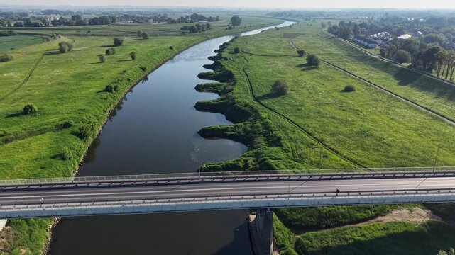 Aerial panoramic view of a car driving across a concrete bridge over a winding river. Scenic summer landscape with green meadows and rural infrastructure.