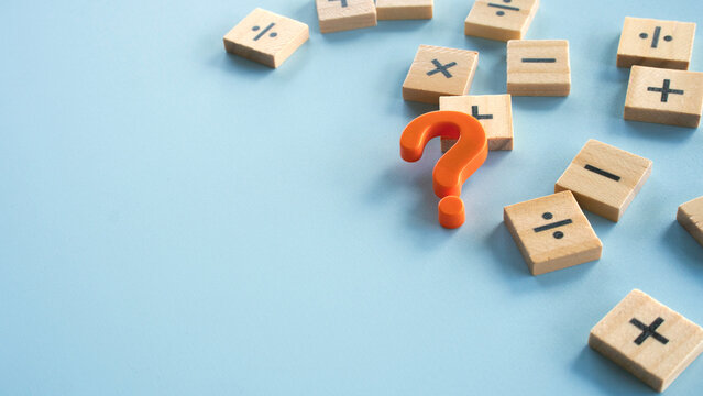 An orange question mark symbol placed amidst scattered wooden tiles with mathematical operator signs, concept of problem solving, confusion, and math questions.