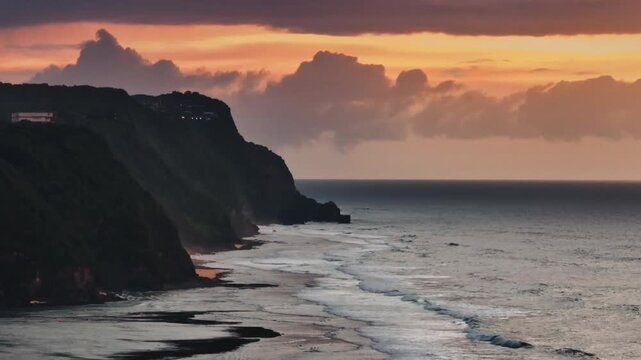 Melasti Beach coastline showing dark cliffs and ocean waves breaking along the shore under a vibrant orange and purple sunset sky with scattered clouds. Nature background. Travel to Bali. Static shot