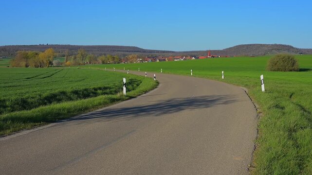 Landstra&szlig;e f&uuml;hrt durch gr&uuml;ne Felder mit Blick auf ein kleines Dorf und blaue Himmel, Ergersheim, Landkreis Neustadt an der Aisch-Bad Windsheim, Mittelfranken, Bayern, Deutschland