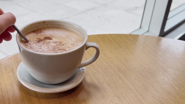 A left-handed person stirring a creamy coffee in a white coffee cup
