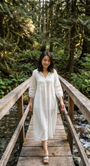Fototapeta premium Smiling young Asian woman walking on a wooden bridge in a lush green forest. Female hiker in white dress exploring nature. Vertical photo